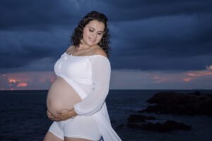 Retrato de maternidad con vestido blanco junto al mar al anochecer